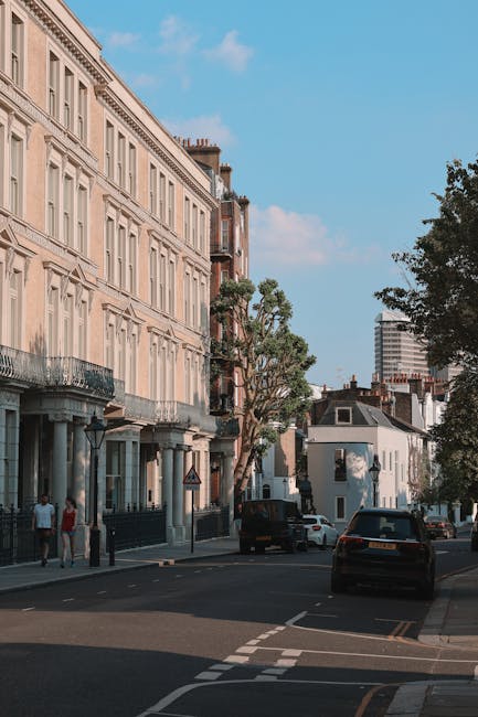 Image of a residential street showing a line of classic terraced houses with beige and cream facades, large windows, and wrought-iron balconies, indicating urban architecture. In the foreground, a bla