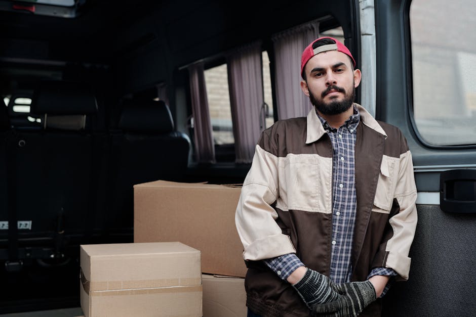 A man with a beard, wearing a black cap, white polo shirt, and jeans, stands inside the open rear of a van loaded with various cardboard boxes of different sizes, some taped and others open, which are
