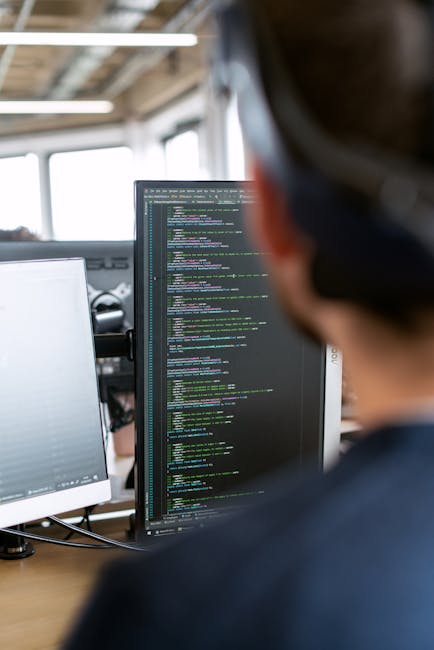 A pair of hands operating a laptop computer on a dark table, showing a screen with code and software development tools. The laptop is positioned indoors, with a black, blurred background. The person's